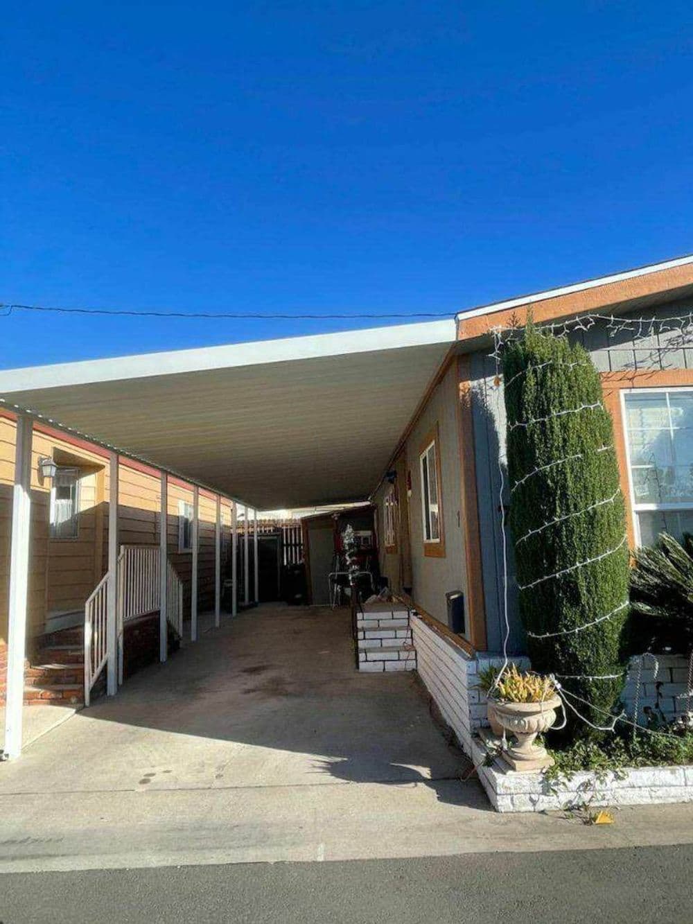 Carport of a mobile home with clear blue sky, plants, and decorative lighting.