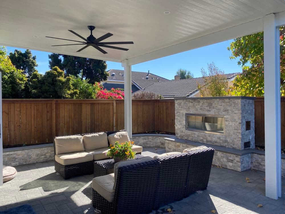 Cozy outdoor patio with seating, fan, and stone fireplace under a shaded roof.