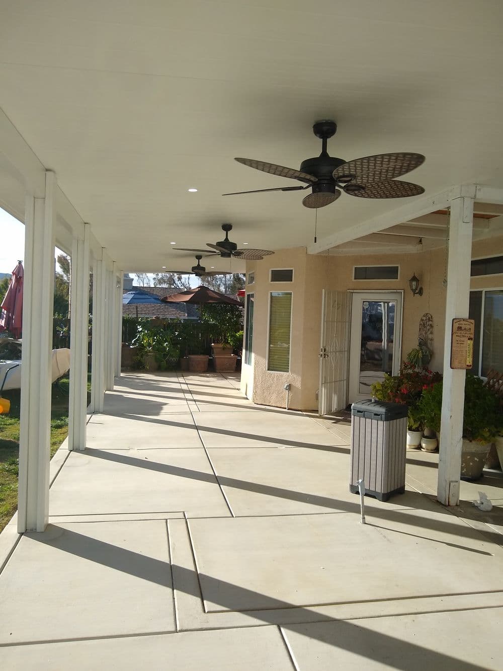 Exterior patio with ceiling fans, concrete flooring, and lush green plants.