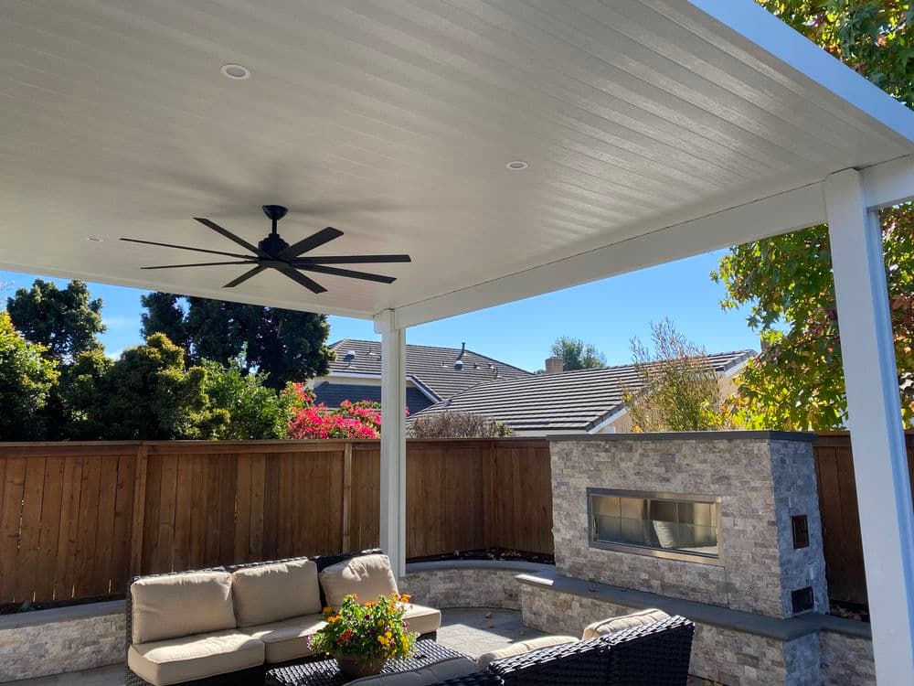 Patio area with a fan, seating, and outdoor stone fireplace under a shaded roof.