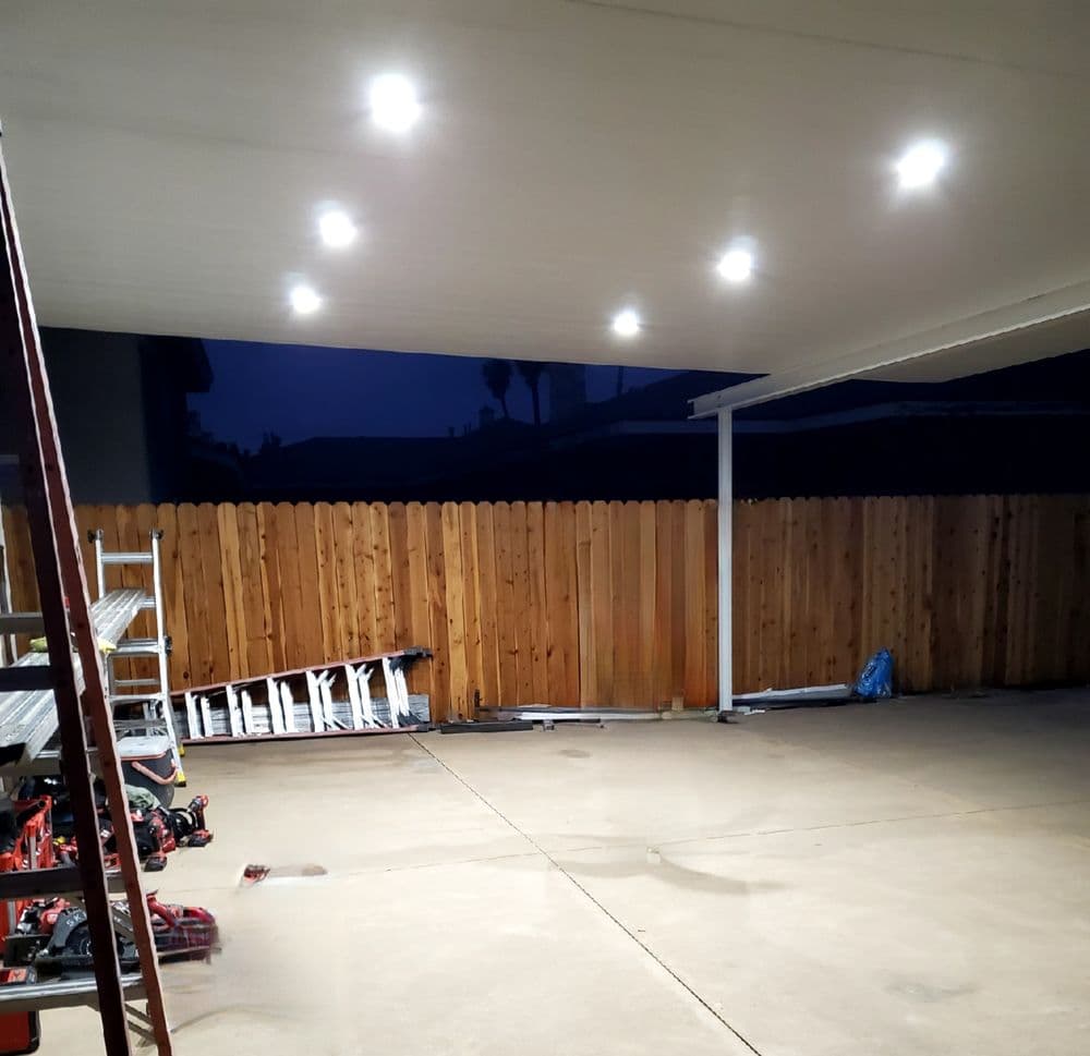 Well-lit patio under a covered awning with wooden fence at dusk, featuring tools and equipment.
