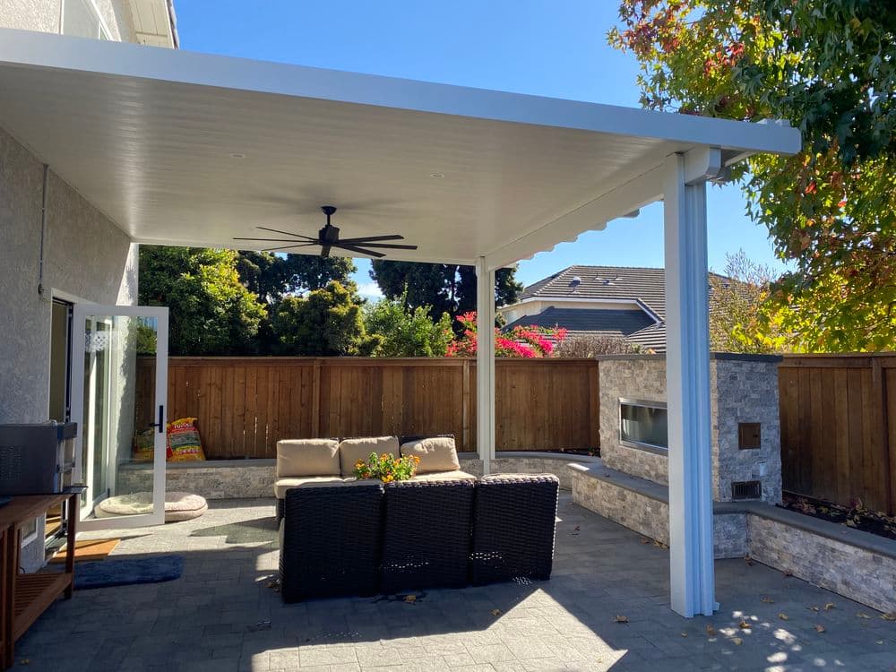 Covered outdoor patio with sofa, fan, and fireplace surrounded by a wooden fence and plants.