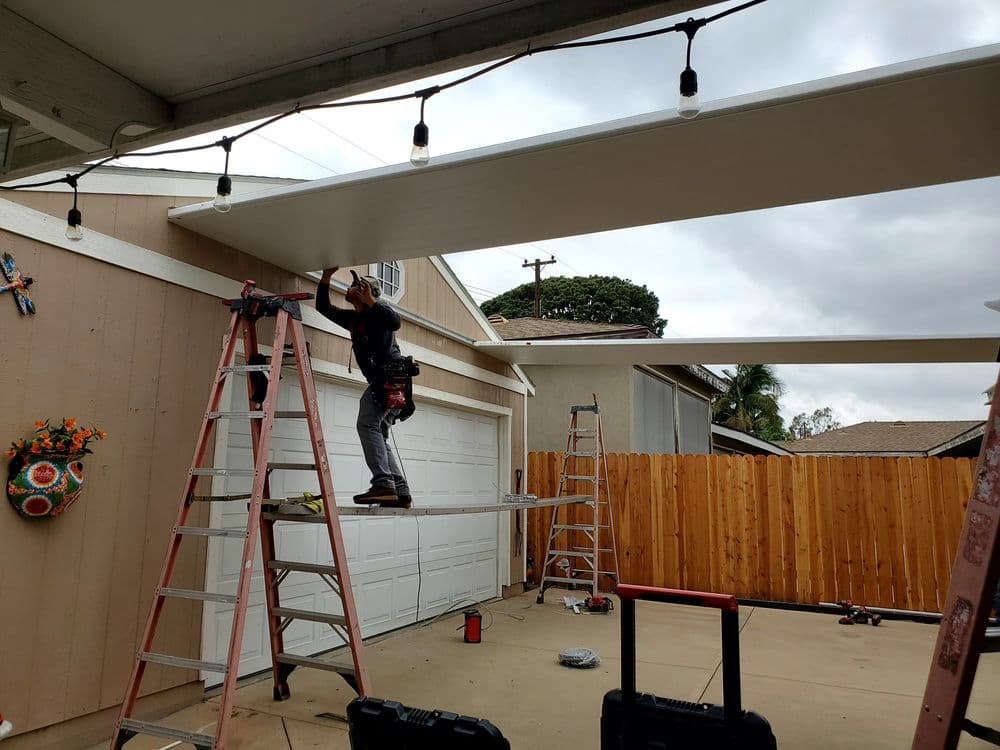 Worker installing a ceiling panel under a patio with string lights in a residential backyard.
