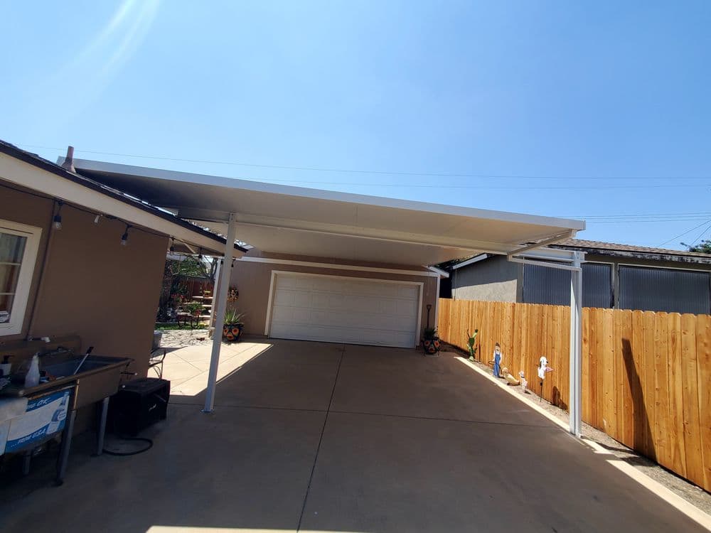 Carport beside a house with a garage and wooden fence under clear blue sky.