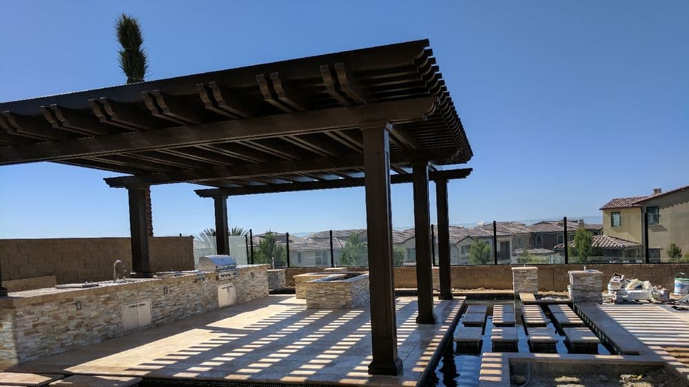 Modern outdoor pergola with kitchen and water features in residential backyard. Bright blue sky.