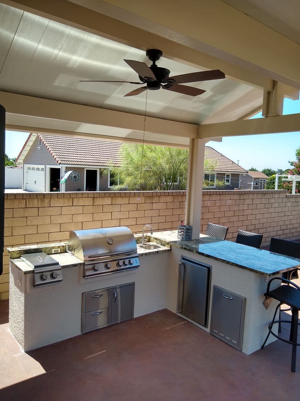 Outdoor kitchen with stainless steel grill, sink, and seating area under a covered patio.